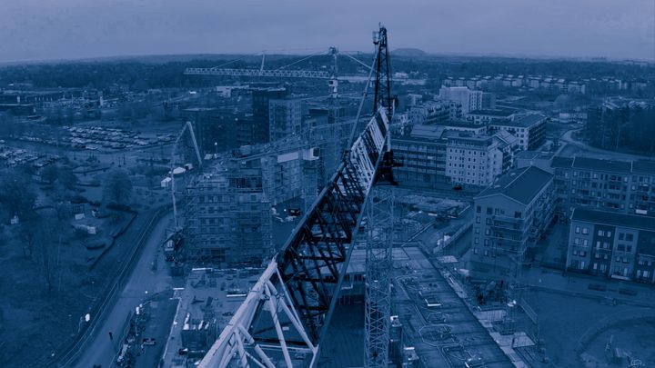 Flygfoto av en byggarbetsplats med en stor byggkran i förgrunden och flera byggnader i bakgrunden.