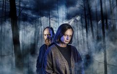 Två personer står i en dimmig skog med berg i bakgrunden.