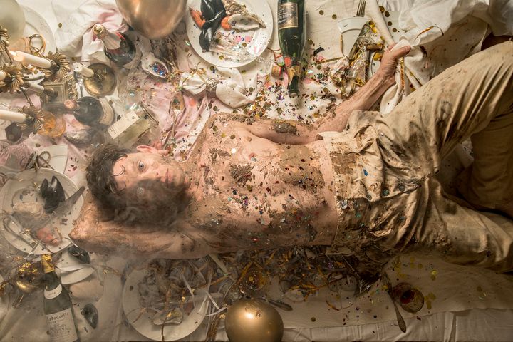 En person ligger på ett golv täckt av konfetti och smuts, omgiven av rester från en fest med flaskor och tallrikar runt sig.