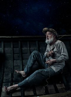 En äldre man med skägg sitter i en liten träbåt under en stjärnklar natt. Han bär en stråhatt och ser upp mot himlen.