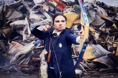 Ung kvinna i mörk kavaj spänner en pilbåge, omgiven av bråte.