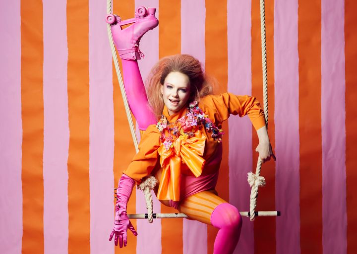 En person klädd i färgglada scenkläder gör en akrobatisk pose på en trapets med en randig bakgrund.