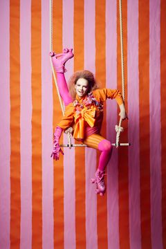 En person i en färgglad, lekfull klädsel sitter på en gunga framför en bakgrund med vertikala rosa och orange ränder.