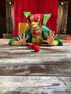 En person utklädd till clown med krona och grön mantel ligger på golvet med röda clownnäsa framför sig.