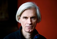 En äldre man i grått hår med seriös min. Röd bakgrund.