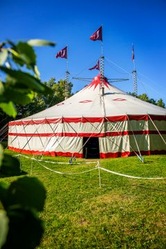 Ett stort cirkustält med röda och vita ränder står på en grön gräsmatta under en klarblå himmel.