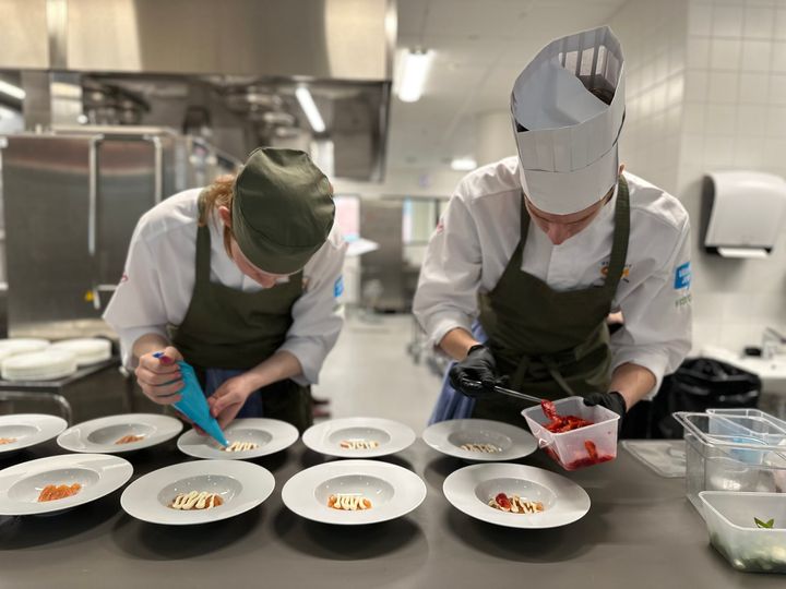 Två kock-elever lägger upp mat på tallrik i ett kök.