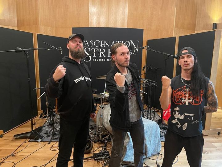 Grupp av tre personer poserar framför musikinstrument i en inspelningsstudio.