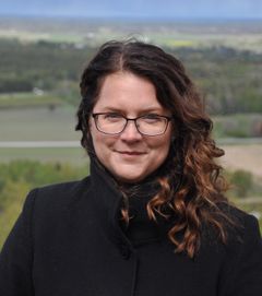 Person med långt lockigt hår och glasögon, klädd i en mörk kappa, står utomhus med landskapet i bakgrunden.