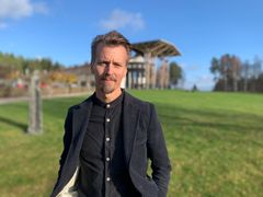 En man i en svart skjorta och jacka som står utomhus på en gräsmatta, med en byggnad i bakgrunden. Det är soligt och himlen är blå.