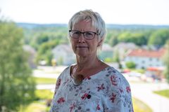 Kvinna med kort blont hår och glasögon står utomhus med suddig bakgrund av hus och träd.