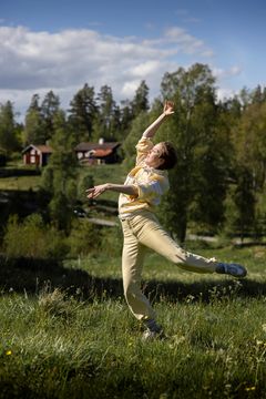 Person dansar på en grön äng med skog och röd stuga i bakgrunden i Tivedstorp.