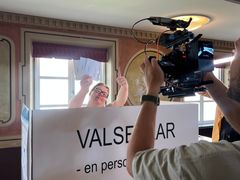 En glad person håller upp valsedlar, han står bakom en stor skylt där det står valsedlar. Personen ser in i en stor videokamera som hålls upp av kamerapersonen i bildens förgrund.