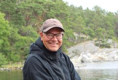 Leende medelålders man med mörka glasögon, beige keps och mörk jacka i grönskande naturlandskap