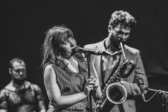 En kvinna sjunger i en mikrofon medan en man spelar saxofon i bakgrunden.