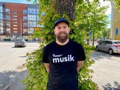 Man med skägg och keps står utomhus. Det står musik på hans svarta t-shirt.