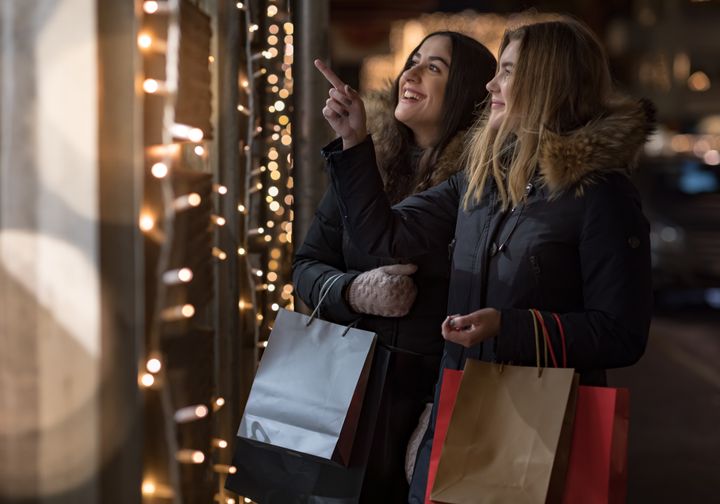 Tvä vänner med shoppingpåsar. Pekar mot ett skyltfönster. Julbelysning i bakgrunden.