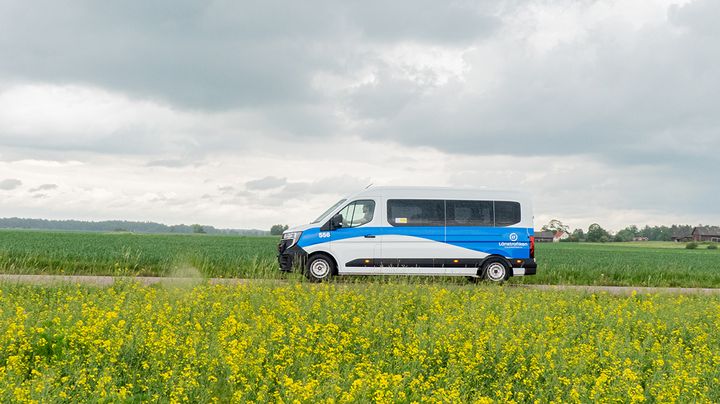 Vitt och blått serviceresefordon som kör på en landsväg med raps i förgrunden.