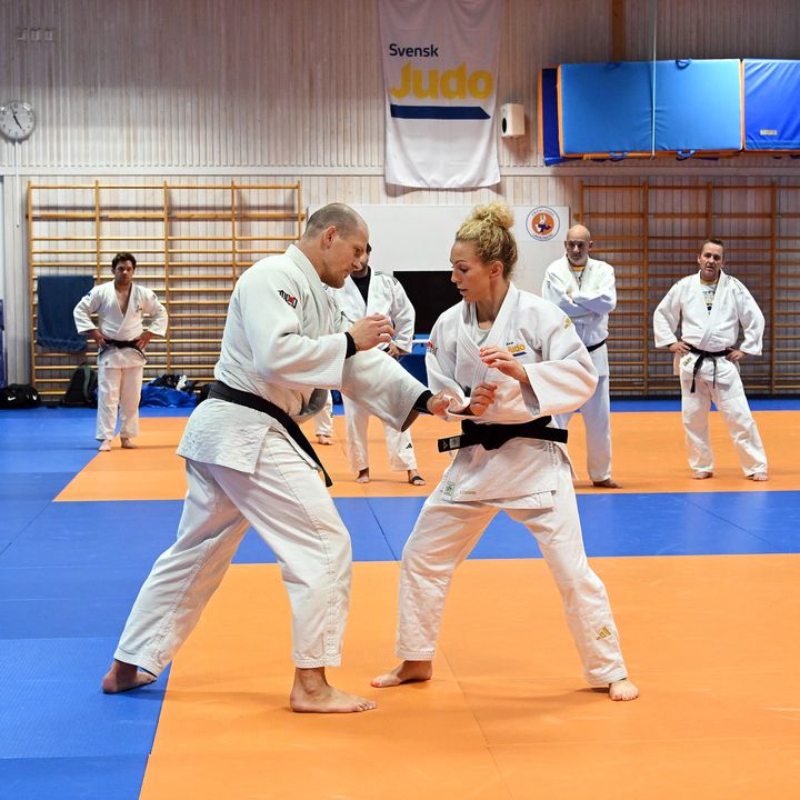 En kvinna och en man i judodräkter övar teknik på en judomatta, med flera personer som tittar på i bakgrunden.