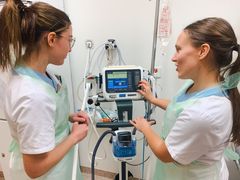 Två sjuksköterskor i vita sjukhuskläder står vid en medicinsk ventilator och diskuterar dess inställningar.