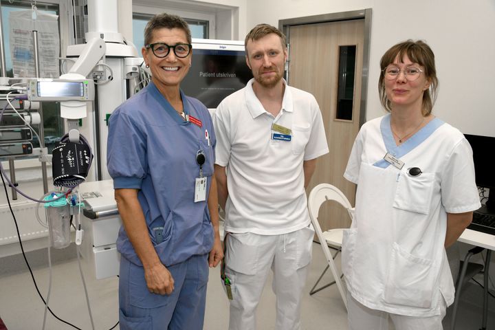 Tre sjukvårdspersonal i arbetskläder står tillsammans i ett sjukhusrum.