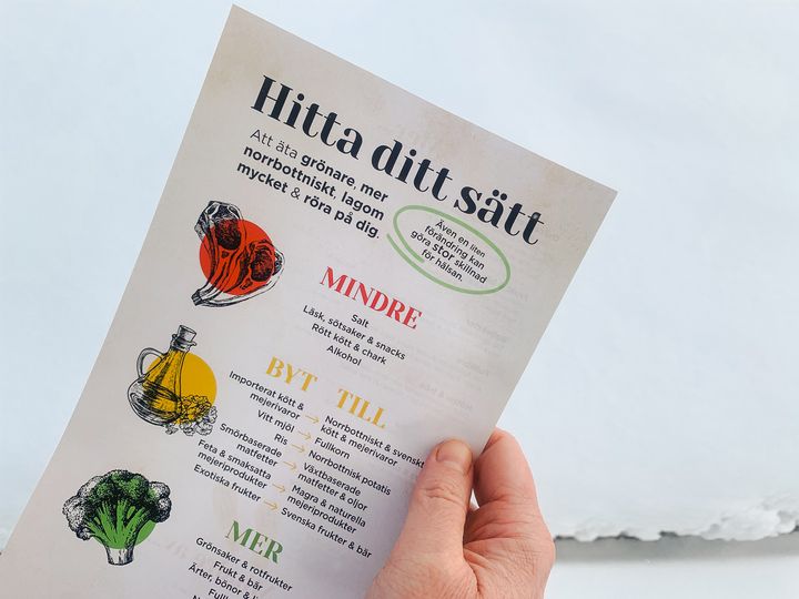 Bild av en hand som håller ett informationsblad med rubriken "Hitta ditt sätt". Bladet innehåller råd om hur man kan äta grönare och mer norrbottniskt, inklusive att äta mindre salt och socker samt byta ut ingredienser. Illustrationer av grönsaker och en flaska olivolja syns också.