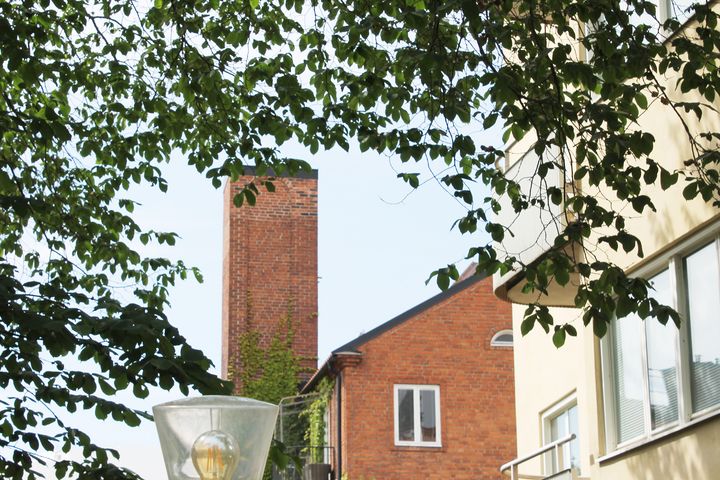 Flerbostadshus och skorsten mot blå himmel och träd i förgrunden.