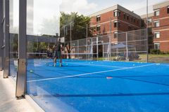 En padelbana med blått underlag och glasväggar, omgiven av träd och en tegelbyggnad i bakgrunden.