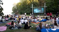 En grupp människor sitter på filtar vid en strand och tittar på en film på en stor utomhusskärm.
