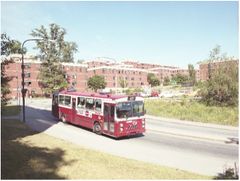 En röd buss åker på Lappis med flerbostadshus i bakgrunden under en solig dag.