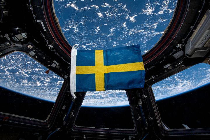 Svenska flaggan svävar i en rymdstation, med jorden synlig genom fönstret i bakgrunden.
