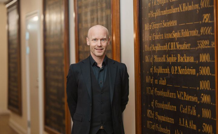 Bild på fastighetschef Mattias i svart kavaj framför en gammal tavla med listor av namn i skrivstil på borgerskapets tidigare medlemmar