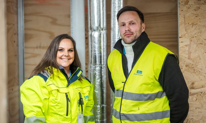 Bild på Veronica och Fredrik framför en vägg iförd varseljackor på en av Einar Mattssons byggarbetsplatser.