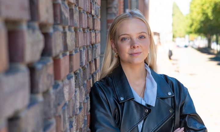 Frida i svart skinnjacka på ljusblå skjorta lutad ute mot en tegelvägg