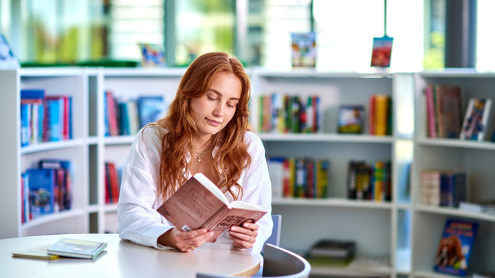 Kvinna sitter vid ett bord i ett bibliotek och läser en bok.