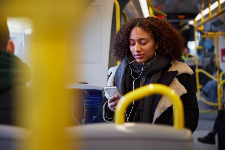 Person som sitter på en buss, lyssnar på musik med hörlurar och håller en mobiltelefon.
