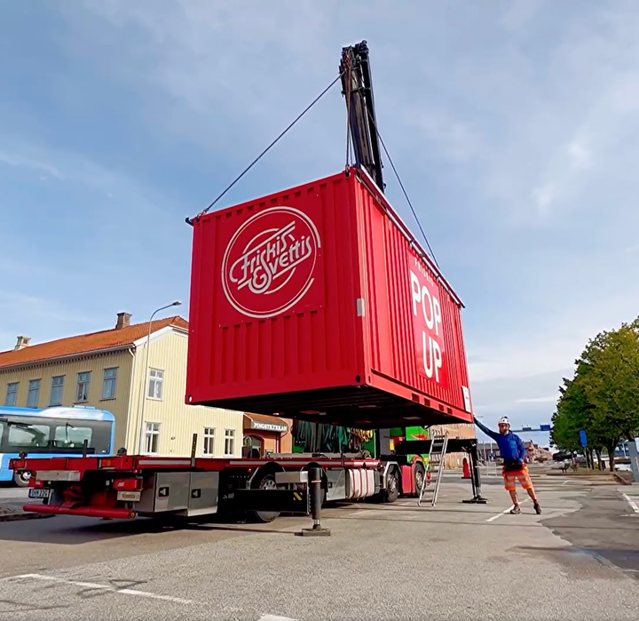 En röd container med texten Friskis&Svettis Pop up lyfts av en kran från en lastbil i en stadsmiljö.