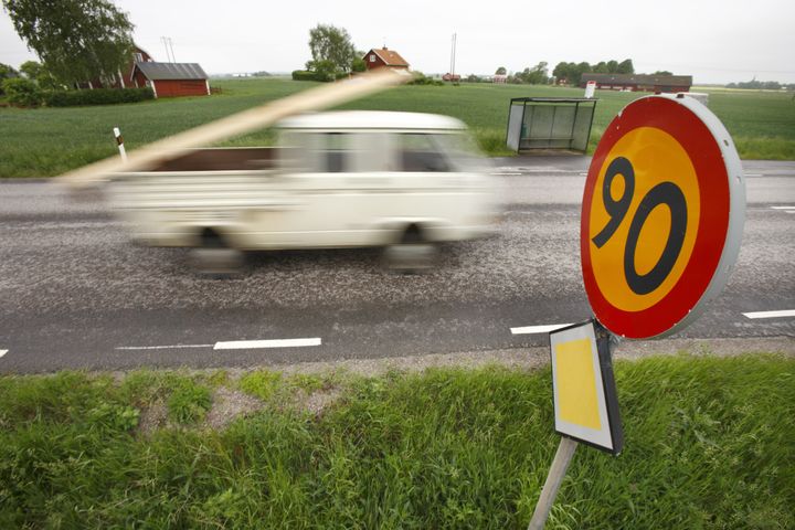 En suddig vit skåpbil som kör förbi en fartgränsskylt med texten "90" på en landsväg.