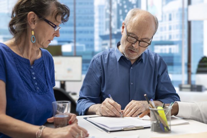 Två personer sitter vid ett skrivbord i ett kontor. En man skriver på ett papper, en kvinna håller ett glas.