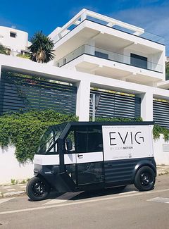 Trehjuligt elfordon med texten "EVIG" parkerat framför en modern vit byggnad.