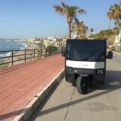 Lastfordonet EVIG kör på en väg vid havet, omgiven av palmer och en kuststad i bakgrunden.