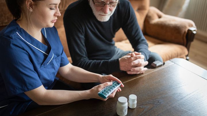 Vårdpersonal sitter i en soffa med en läkemedelsdosett i handen. Hon visar en äldre man med skägg innehållet.