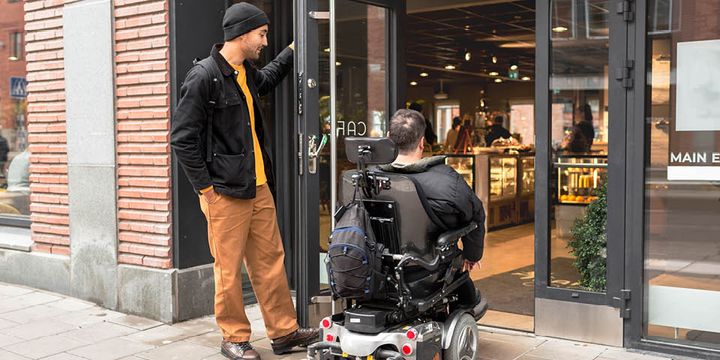 En person står och håller upp en dörr för en person i rullstol.