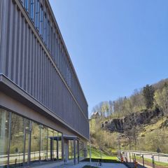 Fasaden på en byggnad med fönsterpartier nedtill och upptill. Intill byggnaden går en väg och en sluttning med berg och skog.