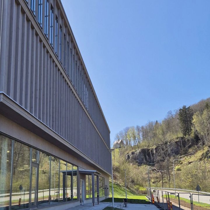 Fasaden%20p%E5%20en%20byggnad%20med%20f%F6nsterpartier%20nedtill%20och%20upptill.%20Intill%20byggnaden%20g%E5r%20en%20v%E4g%20och%20en%20sluttning%20med%20berg%20och%20skog.