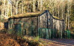 En byggnad i naturfärger som omges av staket i en tät skog.