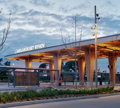 Länssjukhusets nya entréplats sett från sidan. En träkonstruktion i form av tak och pelare med belysning, med texten Länssjukhuset Ryhov. Intill syns en busshållplats och en rad med grön plantering.