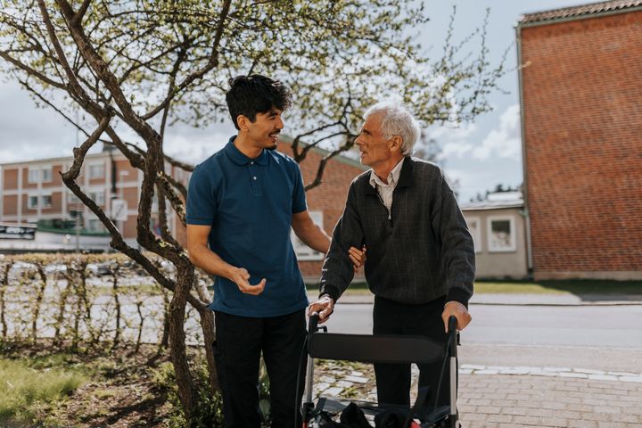 Två personer som står bredvid varandra och tittar på varandra. En yngre och en äldre med rollator.
