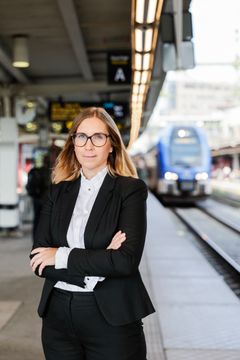 Kvinna i svart kavaj med händer i kors på tågperrong med ett tåg i bakgrunden.