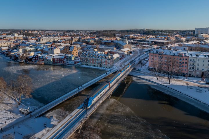 Tåg som i vinterlandskap kör över bro med stad i bakgrunden.